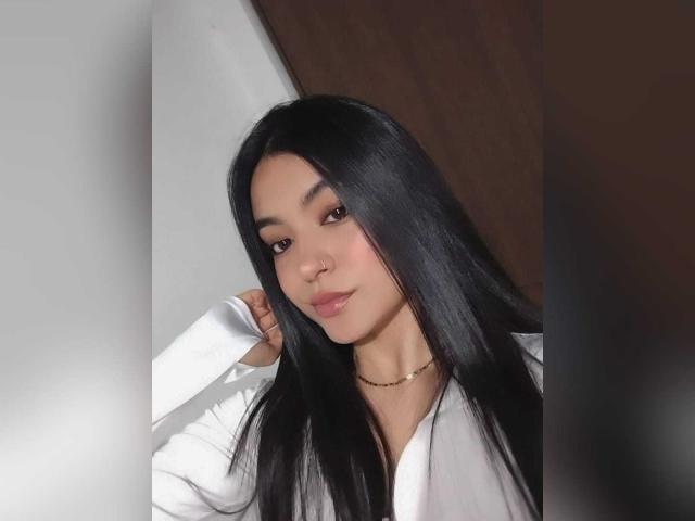 GabriellesRusso with long black hair, wearing a white shirt, has a cute face, subtle makeup, and a gold necklace, looking at the camera with a slight smile.