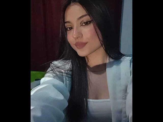 GabriellesRusso, com cabelos longos e pretos, maquiagem leve e batom rosa, vestindo uma blusa branca e um casaco azul claro, tirando uma selfie com um leve sorriso..