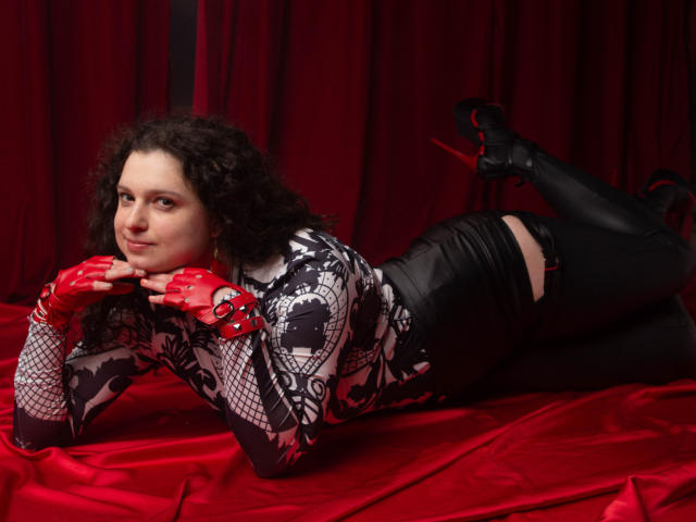 HandmadeMistress has curly brown hair, wearing a black and white patterned top, red fingerless gloves, black thigh-high boots, and black high heels, lying on red satin.
