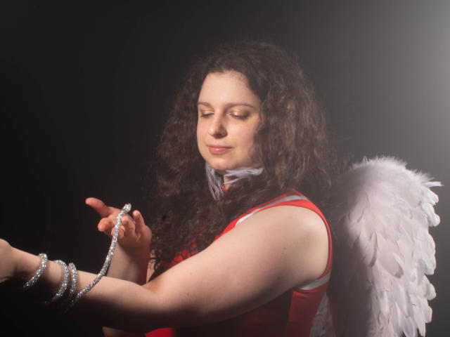 HandmadeMistress with curly brown hair, wearing a red dress and white angel wings, holds a silver bracelet, smiling softly, against a dark background.