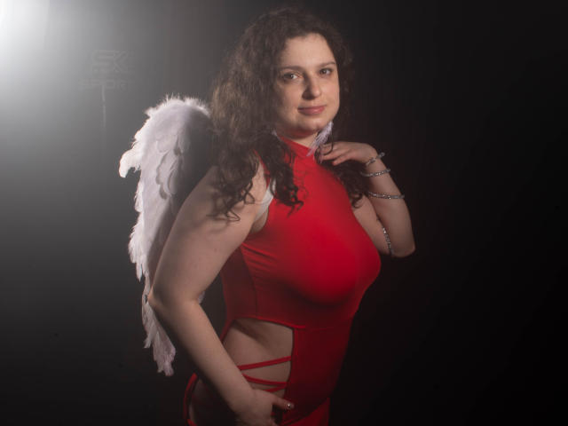 HandmadeMistress with curly brown hair, red dress, and white angel wings, touching her neck, smiling, standing against dark background. Her tits are visible through cut-out dress.