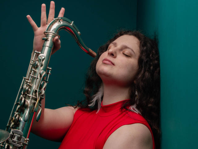 HandmadeMistress with curly dark hair, eyes closed, wearing a red sleeveless top, holding a silver saxophone, leaning against a teal wall, looking blissful.