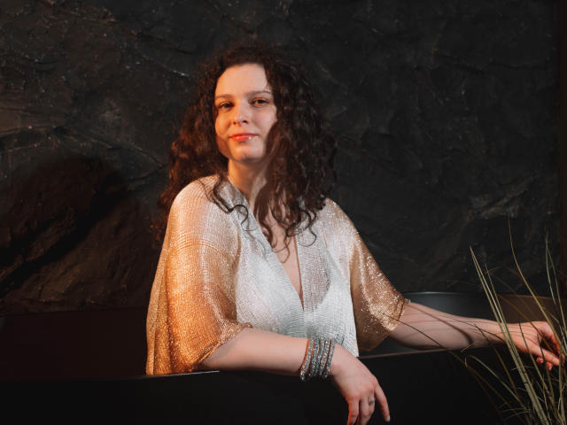 HandmadeMistress with curly brown hair, wearing a shimmering gold and white sheer top, sits by a black surface, her arm resting on it, with a bracelet on her wrist.