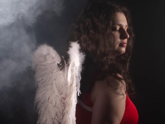 HandmadeMistress with curly brown hair, red top, and white angel wings, side profile, eyes closed, dark background, subtle smile. Boobs visible, soft light, feathers detailed.