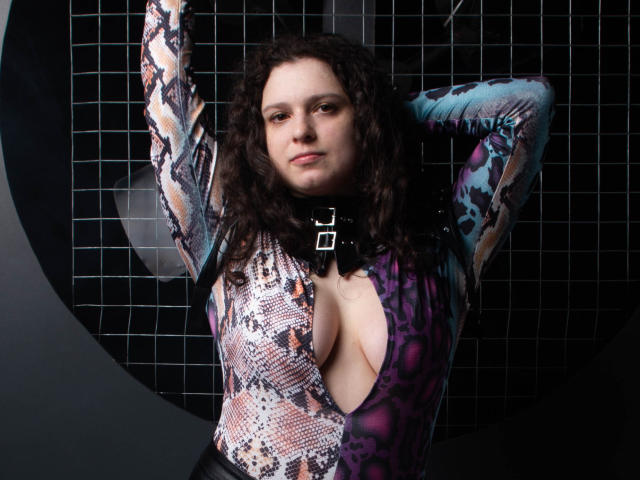 HandmadeMistress with curly brown hair, wearing a snake-patterned, deep-cut, purple and blue leotard, exposing her large, round boobs, and a black choker with a silver buckle.