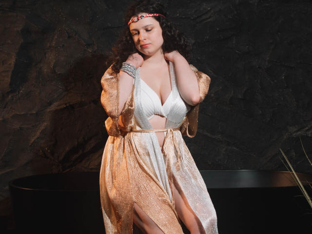 HandmadeMistress with curly brown hair, wearing a shiny gold robe over a white, deep-cut dress showing her big tits and a side thigh slit, standing in a dark room.