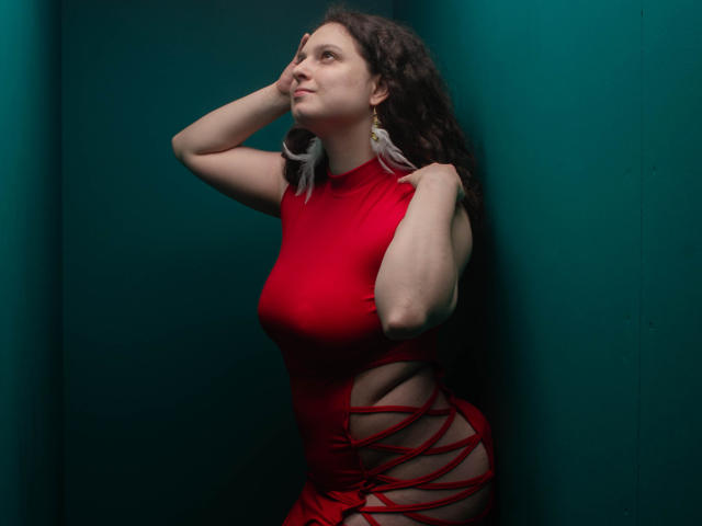HandmadeMistress with curly dark hair, red dress, cutouts on sides, holding her head, looking up, wearing large earrings, standing against teal wall, confident, sexy.