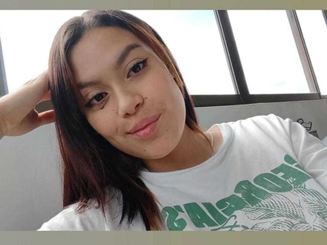 Female-looking person with light brown skin, straight brown hair, small nose, and subtle smile, wearing a white California shirt, hand in hair, window in background, soft light.