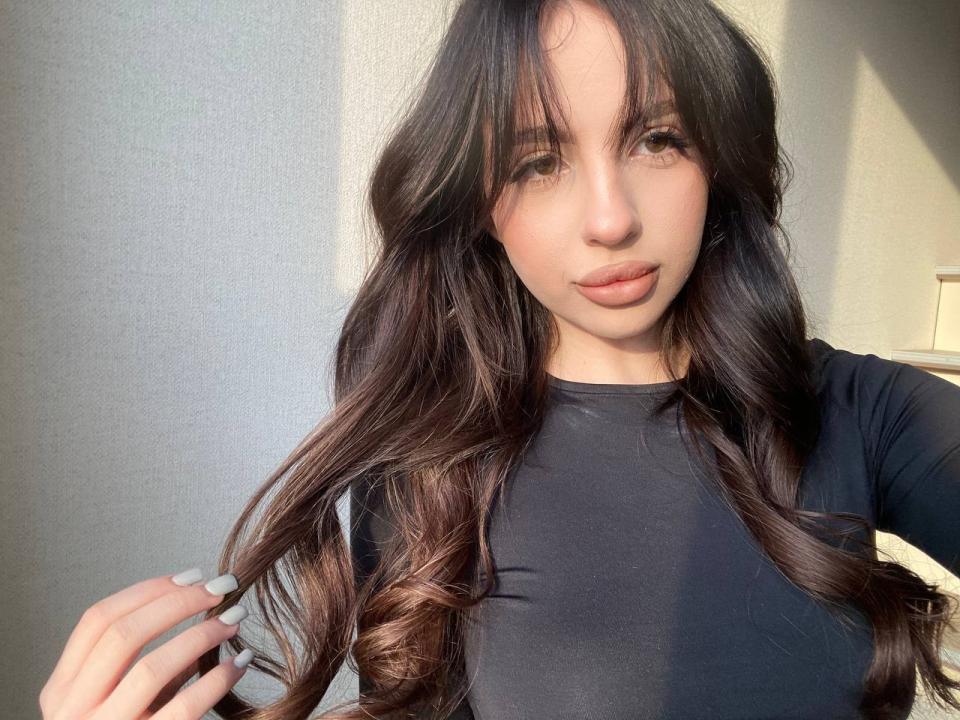 SinfulSapphiree with long, wavy brown hair, light skin, and full lips, wearing a black top, holding her hair with grey nails, sunlight highlighting her face.