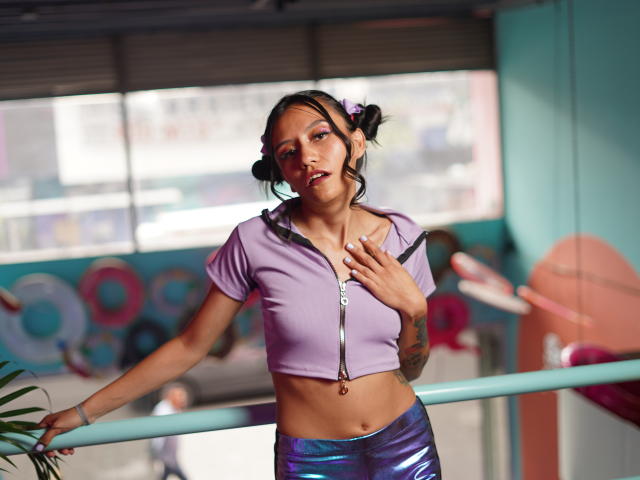 EviieGothel with small tits, wearing a lilac zip-up top and shiny purple pants, has tattoos, dark hair in puffs, and a slight smile, leaning on a ballet barre.