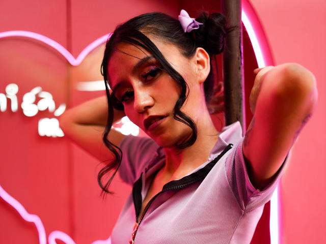 EviieGothel with dark wavy hair in puffy buns, wearing a pink zip-up, has her arms raised, showing her tits and a purple hair clip, in pink neon light.