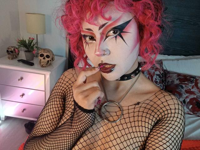 GothVioleth met roze krullend haar, een zwarte top van netstof, donkere make-up, een neusring, een choker en een halsband met ringen, met donkere lipstick op haar tanden, een ring vasthoudend, schedels op een dressoir op de achtergrond..