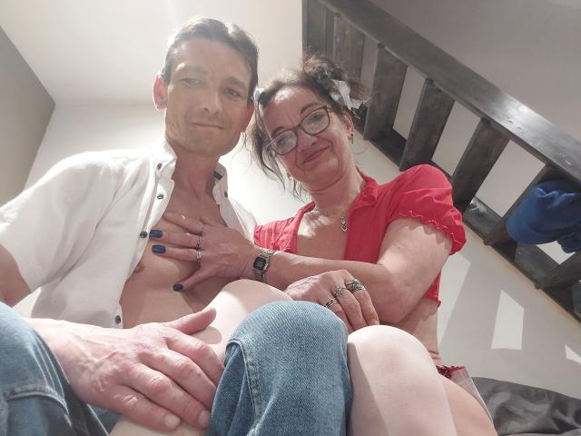 LesCoquinsG (Frau mittleren Alters mit Brille, rotes Hemd) und LesCoquinsG (Mann mittleren Alters, weißes Hemd, keine Hose) sitzen, die Hand der Frau liegt auf der Brust des Mannes, die Hand des Mannes liegt auf ihrem Oberschenkel..