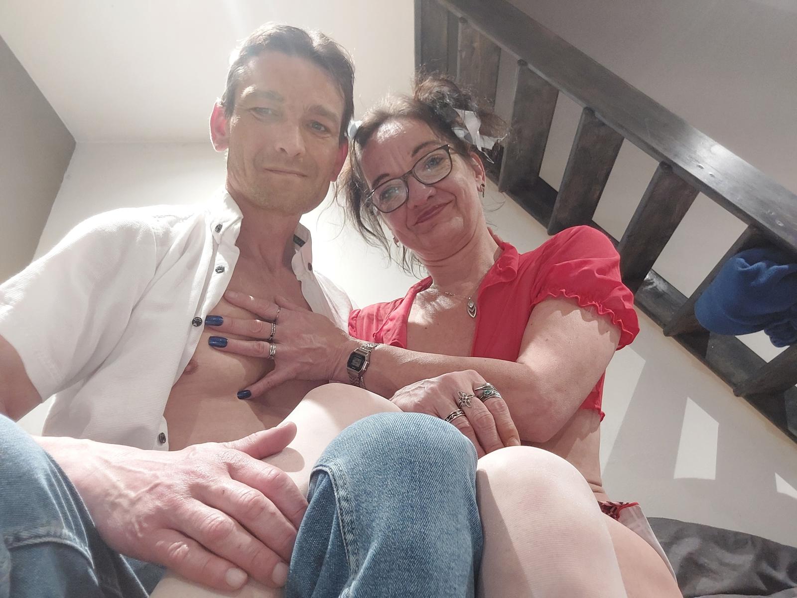 LesCoquinsG (middle-aged woman with glasses, red shirt) and LesCoquinsG (middle-aged man, white shirt, no pants) sitting, woman's hand on man's chest, man's hand on her thigh.