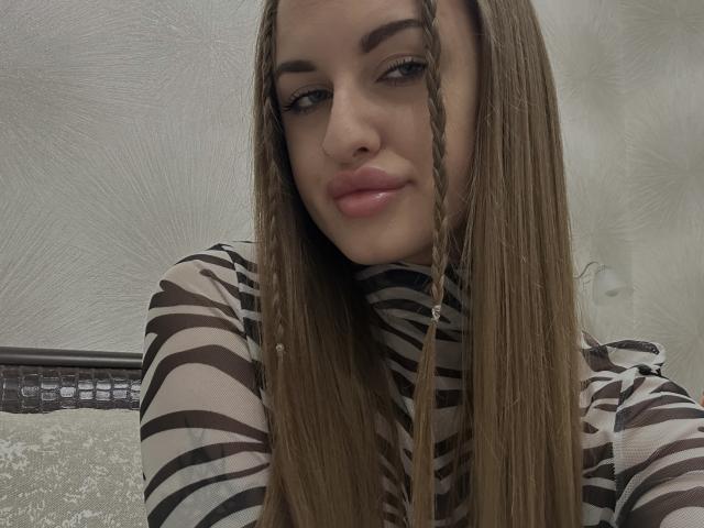 BabeCandy69 with long brown braided hair, wearing a striped brown and beige top, has shiny pink lips and a small nose, looking slightly to the side with a subtle smile.
