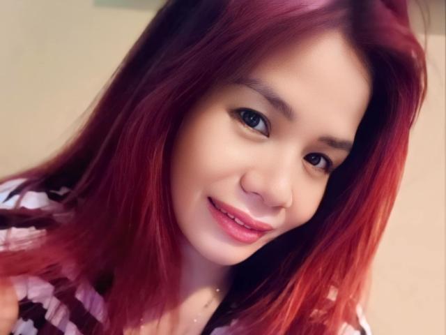 SweetAya69 tiene el pelo rojo, piel clara y está sonriendo con lápiz labial rosa. Lleva una camisa a rayas, y sus senos son ligeramente visibles. Tiene un aspecto lindo y sexy..