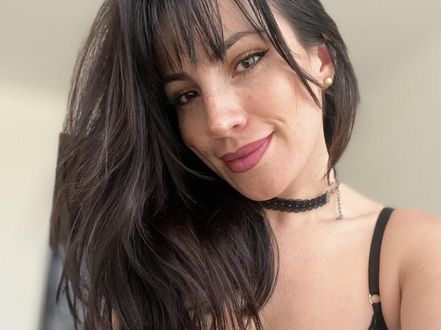 Liliysweet with dark, wavy hair, bangs, and pink lipstick, wearing a black choker and strapless top, smiling at the camera, showing her cute, small tits.