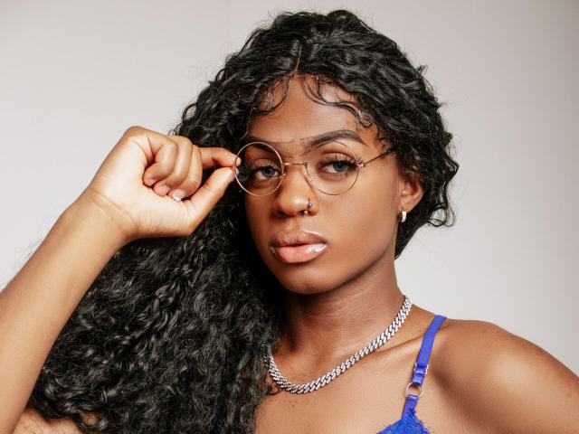 TayssaMaravilhia with curly black hair, wearing gold glasses, nose ring, and blue top, holding glasses with left hand, silver chain necklace, serious expression, medium skin tone.