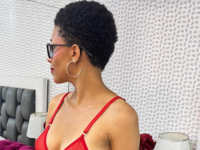 SamantaRoa, con i capelli neri ricci e corti, indossa un reggiseno rosso, grandi orecchini a cerchio, occhiali neri e rossetto rosso, e guarda a sinistra in una camera da letto. Il suo seno di medie dimensioni è visibile..