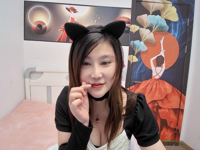 AilsaJade with cat ears, black choker, black top, white cleavage, biting finger, brown hair, pink bed, red dress painting, and floral wallpaper in the background.