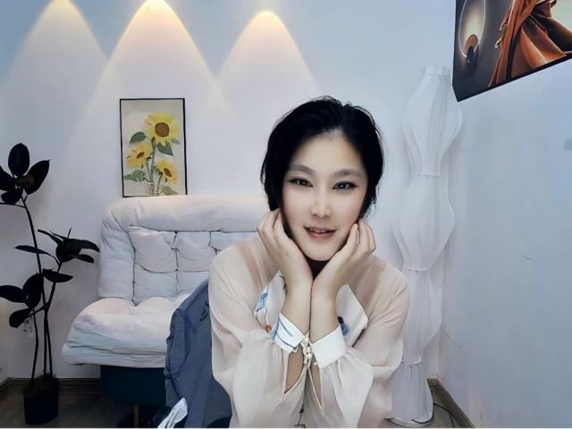 AilsaJade with short black hair, hands on cheeks, smiling, wearing sheer pink top, white blouse, and blue skirt, leaning on white sofa, plant and framed sunflower on wall.
