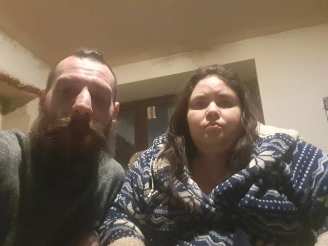 LolaAndAngel with a beard and mustache, wearing a dark green hoodie, next to LolaAndAngel with long brown hair, wearing a blue patterned scarf, pouting.