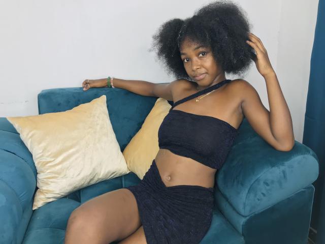 Jaurita with dark skin and curly hair, wearing a black halter top and skirt, sitting on a teal couch with yellow pillows, her hand in her hair, smiling.