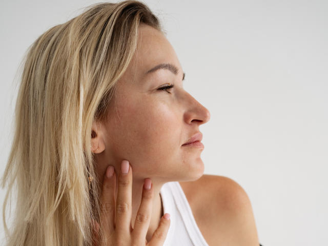 Lilitraya has blonde hair, closed eyes, and a slight smile. She's touching her neck with her right hand, wearing a white tank top. Her face is turned to the right.
