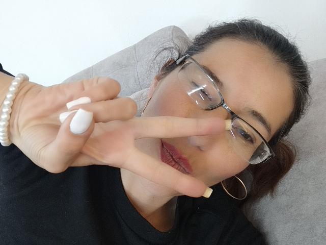 AntonnellaS with glasses, white nails, and pearl bracelet, making a peace sign, wearing a black shirt, and hoop earrings, with red lipstick and relaxed expression.