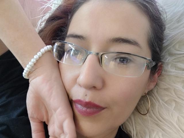 AntonnellaS with glasses, dark hair, red lipstick, and a small nose piercing, lying on a white fluffy pillow, wearing a white bead bracelet and gold hoop earrings.