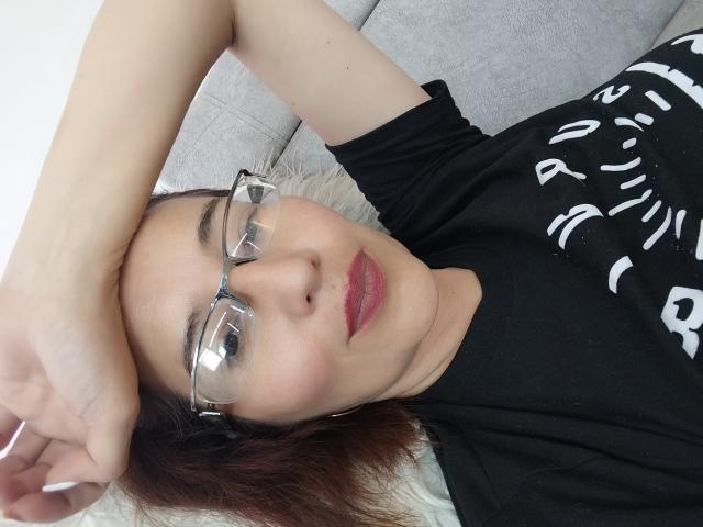 AntonnellaS with glasses, red lipstick, black shirt, and brown hair, lying on a gray couch, arm over head, looking at camera, soft light, relaxed, cute, attractive.