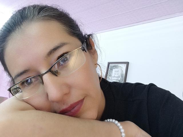 AntonnellaS with glasses, red lipstick, black shirt, white bracelet, resting chin on hand, smiling slightly, brown hair in a bun, pink ceiling, white wall.