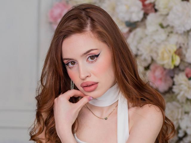 CherylFoxy has long brown hair, pink lips, and white makeup. She's wearing a white scarf, touching her cheek with a hand, and has a necklace. Boobs are not visible.