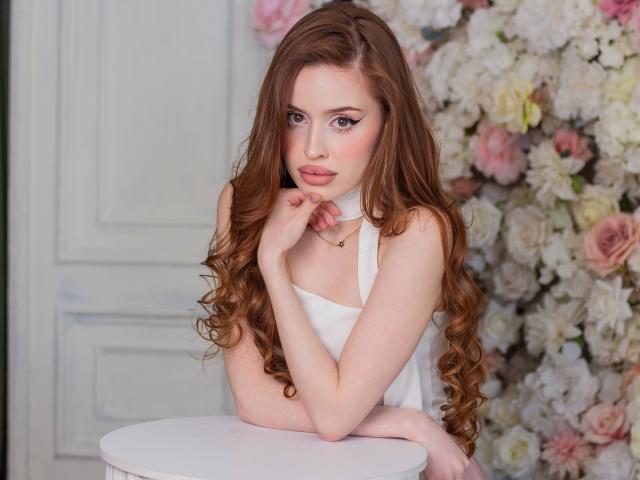 CherylFoxy with long brown curly hair, white sleeveless top, hands near face, pink lips, and blue eyes, sitting at white table, flowers in background, looking cute and sexy.