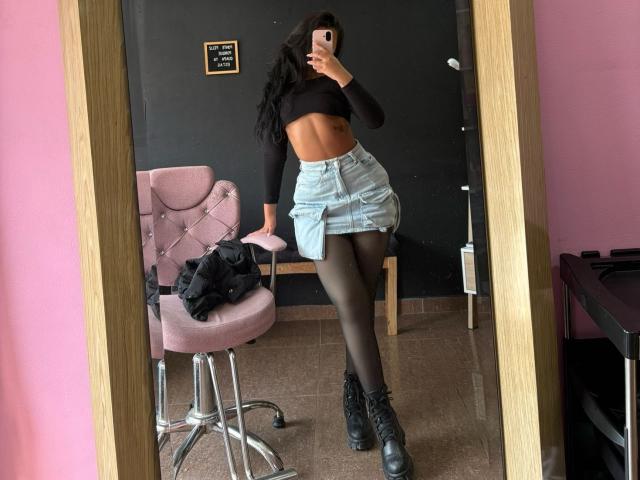 AlessiaFerry in a black crop top, showing her abs, with a blue denim skirt, black tights, and black boots, taking a mirror selfie, standing in a pink room.