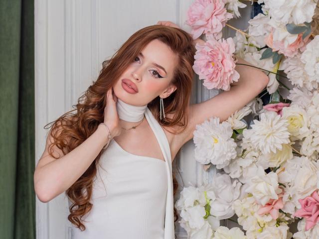 CherylFoxy with long brown hair, wearing a white halter dress, touching her neck, surrounded by pink and white flowers, looking at the camera with pouty lips.