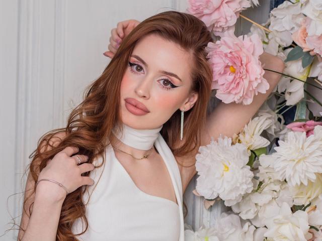 CherylFoxy with long red hair, pink eyeshadow, white halter top, earrings, and a necklace, arms raised, surrounded by pink and white flowers, hands touching her hair.