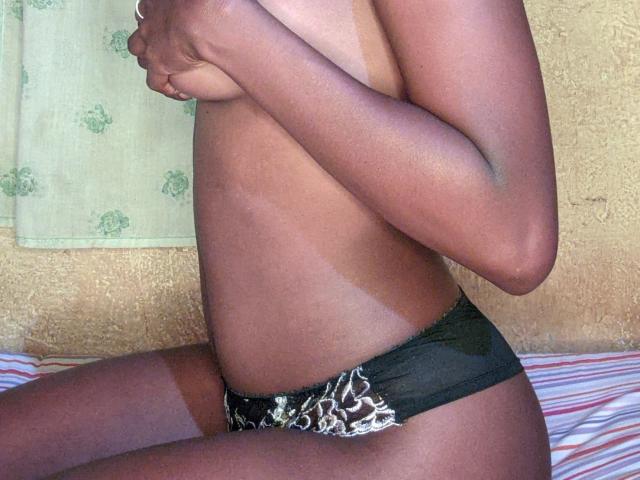 MilaLove69's tan-lined, dark-skinned body, topless with hands covering her tits, wearing black leopard-print panties, sitting on a striped bed.