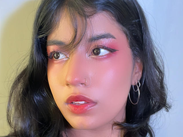 LunaCooC hat dunkles, welliges Haar, rosigen Lidschatten und roten Lippenstift. Sie trägt Ohrringe im Stecker-Stil, eine Nasenring und ihre glänzenden, rosafarbenen Lippen sind leicht geöffnet..