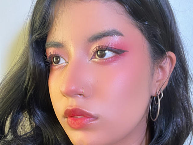 Lunas Gesicht, mit glänzendem rosafarbenem Lidschatten, schwarzem Eyeliner, Nasenring, glänzlich roten Lippen, schwarzen, welligen Haaren und Reifenohrringen, blickt leicht zur Seite..