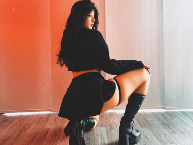LunaCooC mit langem, dunklem Haar, hockt, trägt ein schwarzes Crop-Top, einen Minirock, schwarze, hohe Stiefel, zeigt ihr Gesäß, vor rotem, orangefarbenem Licht. Ihr Hintern ist in der Luft..