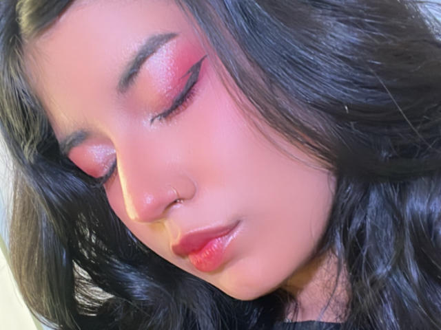 A LunaCooC tem pálpebras rosa brilhantes, cabelo ondulado escuro, um piercing no nariz, lábios vermelhos brilhantes e olhos fechados, parecendo serena com um leve sorriso. Suas nádegas não estão visíveis..
