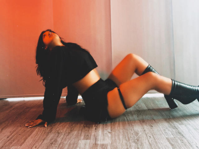 LunaCooC liegt auf dem Boden, den Rücken gekrümmt, trägt ein schwarzes Crop-Top und Shorts, eine Strumpfhose am Oberschenkel und kniehohe Stiefel, wobei rotes Licht einen warmen Schein erzeugt..