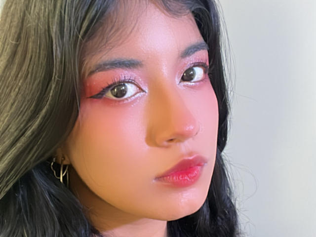 Lunas Gesicht, mit glänzendem pinkfarbenem Lippenstift, rotem Lidschatten und langen schwarzen Haaren, zeigt ein leichtes Lächeln. Sie trägt einen Nasenring und goldene Ohrringe..