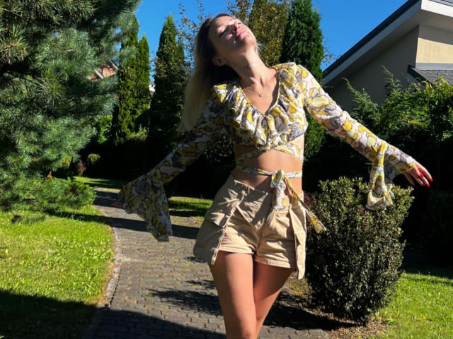 Lilitraya has tits covered by a yellow floral crop top, tan shorts with a tied waist, and is dancing in sunlight with eyes closed and head tilted back.