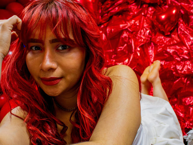 MelannyTaylorr with red curly hair, red lipstick, and a necklace, lying on her stomach, showing her bare shoulders and white dress, with her feet up, smiling at the camera.