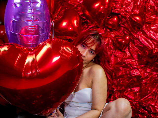 MelannyTaylorr with red hair, wearing a white strapless top, holding a shiny red heart balloon, surrounded by red balloons, and a purple balloon, in a colorful, festive setting.