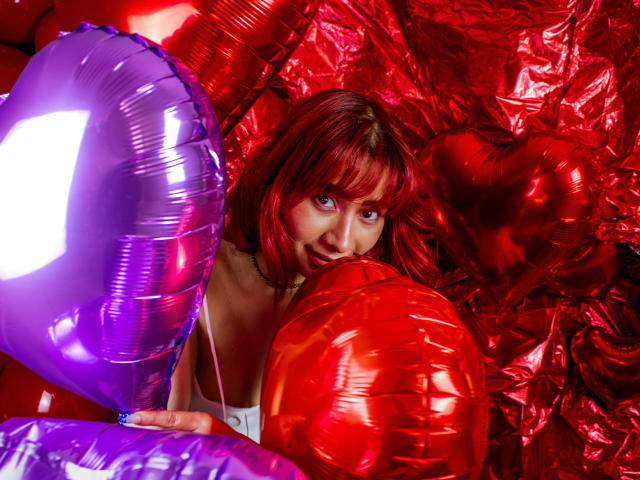 MelannyTaylorr with red hair, holding purple and red shiny balloons, looking up with a playful smile, her tits slightly visible under a white top.
