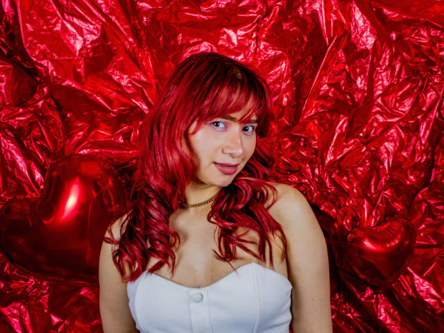 MelannyTaylorr with red hair, blue eyes, white strapless top, small tits, fair skin, gold necklace, smiling, red crinkled background, shiny red heart balloons on the left.