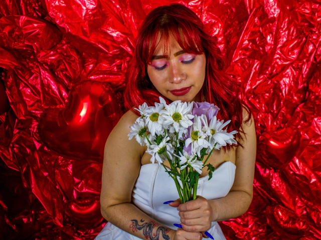 MelannyTaylorr with red hair, pink eyeshadow, holding white daisies, wearing white strapless dress, blue nails, tattoo on right wrist, red shiny background, heart-shaped object to the left.